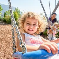 Ein lachendes Kind schaukelt im Vordergrund auf einem Spielplatz, umgeben von sommerlicher Natur und Bergen im Hintergrund. Im Hintergrund schaukeln weitere Kinder mit einer Betreuungsperson. Die Szene strahlt Freude, Unbeschwertheit und gemeinsames Spiel an der frischen Luft aus.