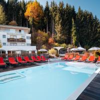 Moderner Außenpool mit hellblauem Wasser, umgeben von roten Liegestühlen und weißen Sonnenschirmen vor einem stilvollen Alpenhotel. Die lebhaften Herbstfarben der umgebenden Bäume sorgen für eine angenehme Wohlfühl-Atmosphäre und laden zum Entspannen ein. Ideal für erholsame Momente inmitten der Natur.