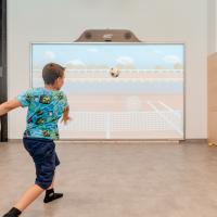 Ein Junge spielt Fußball an einer interaktiven Medienwand in einem Indoor-Spielbereich. Die Projektionsfläche zeigt ein virtuelles Spielfeld, das zum aktiven Mitmachen einlädt. Helle, freundliche Umgebung mit Kletterwand im Hintergrund und viel Bewegungsfreiheit für Kinder.
