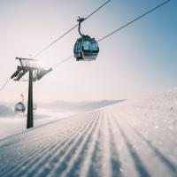 Eine moderne Seilbahn fährt bei strahlendem Sonnenschein über eine frisch präparierte Skipiste. Der glitzernde Schnee und die klaren Himmel vermitteln eine ruhige Winteratmosphäre, ideal für Wintersportler in den Alpen. Die Bildkomposition betont Tiefe und Weite der verschneiten Landschaft.