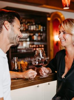 Ein entspanntes Paar genießt ein Glas Rotwein an der stilvoll eingerichteten Bar. Im Hintergrund sind beleuchtete Flaschenregale sichtbar, die eine gemütliche und einladende Atmosphäre erzeugen. Die beiden Personen lächeln sich an und wirken glücklich, was dem Bild eine warme Stimmung verleiht.