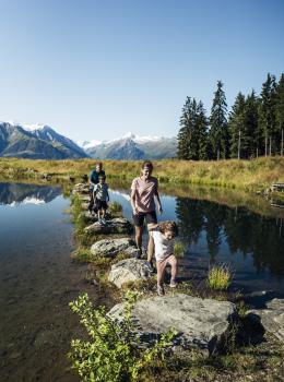 Eine Familie überquert gemeinsam einen idyllischen Bergsee auf Trittsteinen. Im Hintergrund erheben sich majestätische Alpen mit verschneiten Gipfeln und dichten Wäldern. Das Sonnenlicht spiegelt sich im klaren Wasser, während die Natur eine entspannte Urlaubsatmosphäre bietet.