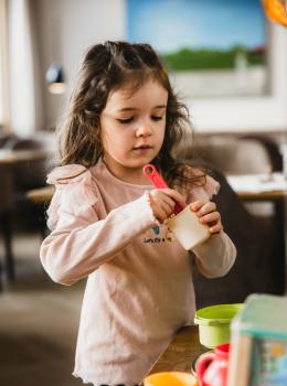Ein junges Mädchen steht an einem Tisch und spielt konzentriert mit buntem Spielgeschirr. Sie hält einen kleinen Messlöffel in der Hand und taucht ihn in einen Becher. Im Hintergrund ist ein gemütlicher, heller Raum mit modernen Möbeln sichtbar. Die Szene vermittelt eine freundliche und kreative Atmosphäre.