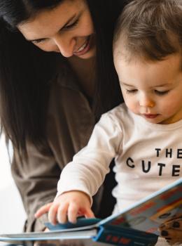 Eine Mutter und ihr Kleinkind genießen gemeinsam eine liebevolle Vorlesestunde. Das Kind zeigt neugierig auf ein buntes Bilderbuch, während die Mutter lächelnd zusieht. Die Szene vermittelt Geborgenheit, Nähe und Freude am gemeinsamen Entdecken von Geschichten.