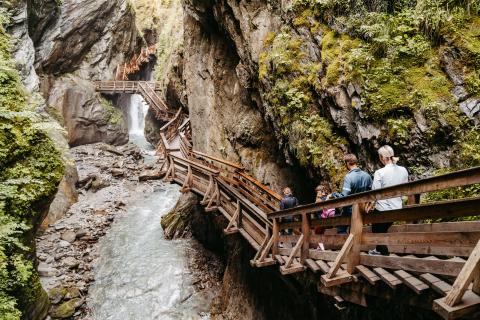 Eine Gruppe Spaziergänger erkundet einen schmalen Holzsteg entlang einer felsigen Klamm mit tosendem Wasserfall im Hintergrund. Üppiges Grün wächst an den steilen Felswänden und verleiht der malerischen Schlucht eine natürliche, abenteuerliche Atmosphäre.