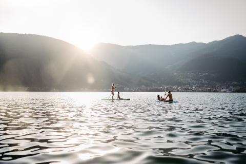 Zwei Personen genießen Stand-Up-Paddling auf einem ruhigen See vor der Kulisse sanfter Berge bei Sonnenuntergang. Die Sonnenstrahlen spiegeln sich im Wasser, während im Hintergrund sanfte Hügel und ein kleines Dorf zu sehen sind. Eine entspannte, naturnahe Atmosphäre.