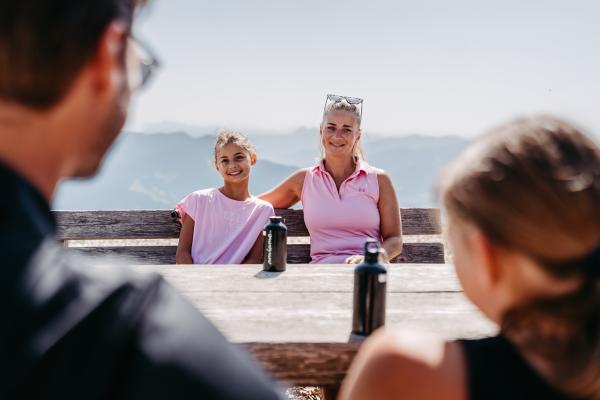 Eine lachende Mutter mit ihrem Kind genießt den Tag auf einer Berghütte, während Vater und Tochter ihnen gegenüber sitzen. Im Hintergrund erstreckt sich eine beeindruckende Alpenlandschaft. Wasserflaschen auf dem Holztisch runden die gemütliche Stimmung ab.