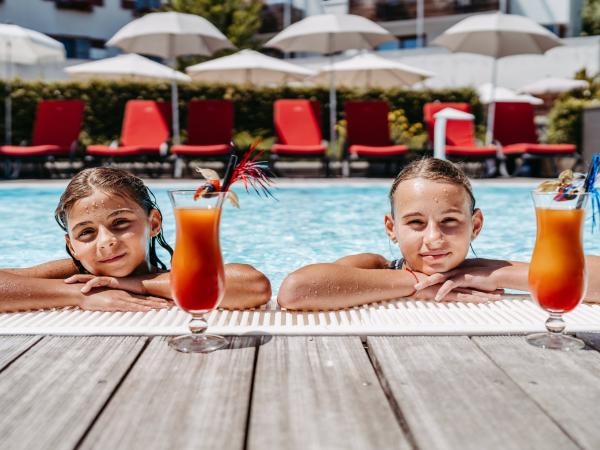 Zwei Kinder genießen einen sonnigen Tag im Freibad und lehnen entspannt am Beckenrand. Vor ihnen stehen bunte, erfrischende Cocktails mit Deko, im Hintergrund sorgen rote Liegen und weiße Sonnenschirme für Urlaubsstimmung. Die Szene vermittelt Sommer, Erholung und Lebensfreude.