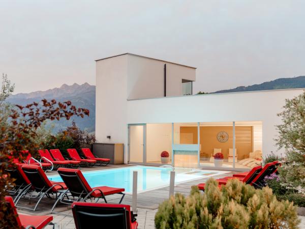 Ein stilvoll gestalteter Außenpool mit eleganten roten Liegestühlen lädt zum Entspannen ein. Im Hintergrund beeindruckt die alpine Berglandschaft, während das moderne Gebäude mit großen Glasfronten für eine helle und einladende Atmosphäre sorgt. Ideal für erholsame Stunden im Freien.