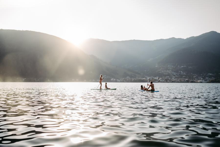 Zwei Personen genießen Stand-Up-Paddling auf einem ruhigen See vor der Kulisse sanfter Berge bei Sonnenuntergang. Die Sonnenstrahlen spiegeln sich im Wasser, während im Hintergrund sanfte Hügel und ein kleines Dorf zu sehen sind. Eine entspannte, naturnahe Atmosphäre.